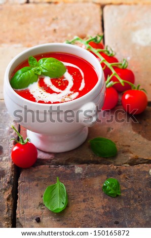 Small bowl of colourful red tomato soup with cream and basil on old weathered bricks with scattered fresh ingredients