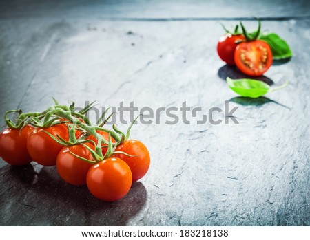 Fresh grape tomatoes on the vine in the lower corner of a dark textured wooden surface with highlight and vignetting and a halved tomato in the top corner with copyspace between