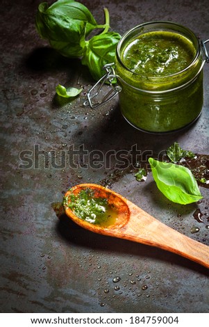 Jar of fresh pesto with basil being prepared in a country kitchen with olive oil in a wooden spoon on an old textured slate surface
