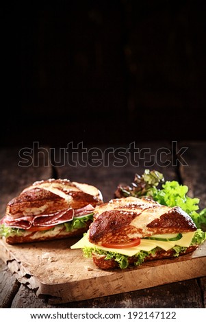 German lye rolls, glazed with a lye solution before baking, filled with cheese and spicy salami sausage with fresh crispy lettuce, cucumber and tomato served on a rustic wooden board with copyspace
