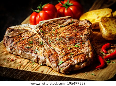 Grilled T-bone steak seasoned with spices and fresh herbs served on a wooden board with fresh tomato , roast potatoes and red hot chili peppers