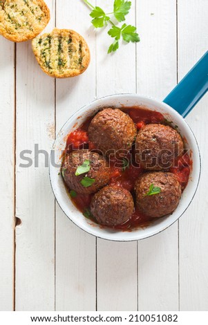 Tasty savory ground beef meatballs in tomato sauce served in a saucepan with crusty toasted herb bread on a white rustic wooden table, overhead view