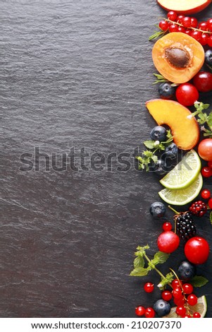 Border of fresh fruit and herbs along the right of the frame with assorted berries, apricot, peach and lemon on a dark grey slate background with copyspace