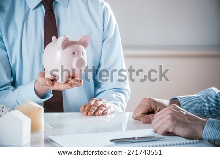 Businessmen discussing house costs and funding with a client holding a piggy bank alongside insulation samples while the engineer or architect takes notes in a savings and achievement concept