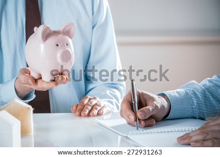 Close Up of Business Man Holding Pink Piggy Bank in Meeting with Co-Worker Taking Notes in Note Book, Real Estate or Investment Concept Image