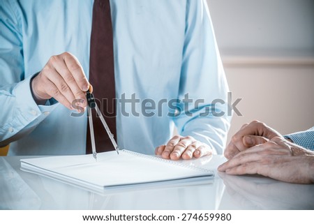 Male engineer wearing blue shirt and tie while measuring with a compass tool a technical drawing at his desk together with a colleague
