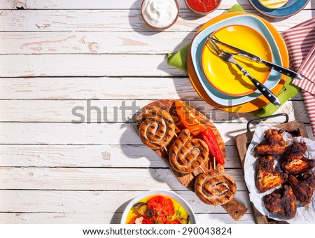High Angle View of Grilled Sausages and Chicken Wings on Picnic Table with Colorful Plates and Cutlery, Copy Space on Table with Barbequed Meal