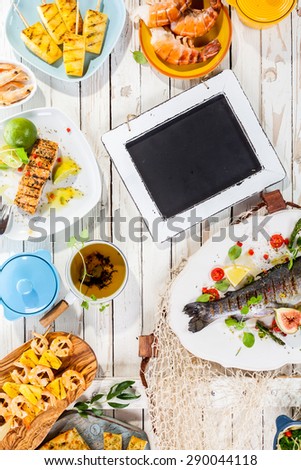 Small Chalkboard on Rustic White Wooden Table Surrounded by Grilled Seafood and Fruit Dishes