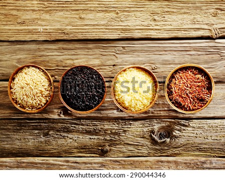 Four different varieties of dried rice, wild, basmati, long-grain and red, displayed in individual wooden bowls in a line on an old rustic weathered wooden table, overhead view with copyspace