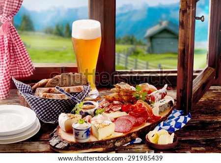 Bavarian tavern lunch with bread, assorted cold spicy sausage and meat, cheese and a long ice cold beer on a rustic wooden windowsill with a view of the Alps