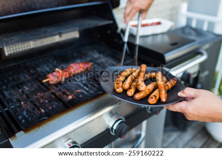 BBQ with sausages and red meat on the grill - male hands holding a plate and taking the meat off the grill before it is too late