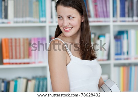 pretty female college student in the university library, carrying books in front of bookshelves (color toned image)