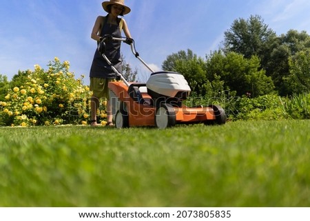 A young girl is mowing a lawn in the backyard with an orange lawn mower. A woman gardener is trimming grass with the grass cutter, bottom view.A lawnmower is cutting a lawn on a summer sunny day.