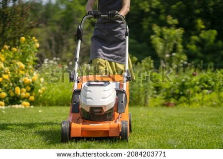 A young girl is mowing a lawn in the backyard with an orange lawn mower. A woman gardener is trimming grass with the grass cutter. A lawnmower is cutting a lawn on a summer sunny day. Foto stock © 