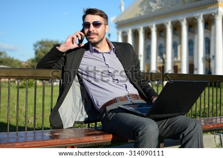 Bearded man in sunglasses with a laptop is sitting on a bench and speaking on the phone