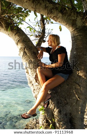 A woman is looking over the ocean while sitting in a large tree.