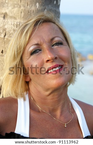 A woman in her 40\'s is looking at the viewer while leaning on a palm tree at the beach.