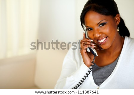 Portrait of a smiling black woman looking at you while talking on phone at home indoor. With copyspace