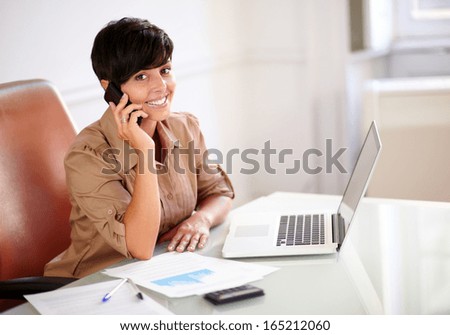 Portrait of an entrepreneur lady speaking on her cellphone while looking at you and sitting on office desk