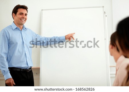 Portrait of a latin adult businessman on blue shirt pointing at whiteboard while smiling and standing in front of colleagues on workplace - copyspace