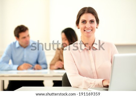 Portrait of lovely professional woman smiling at you while working on her laptop sitting in front of two coworker on office desk