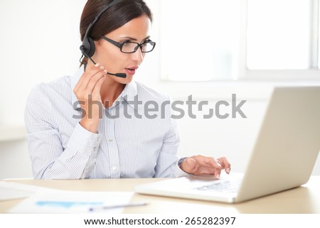 Pretty latin employee with spectacles talking on her headphones while using the computer
