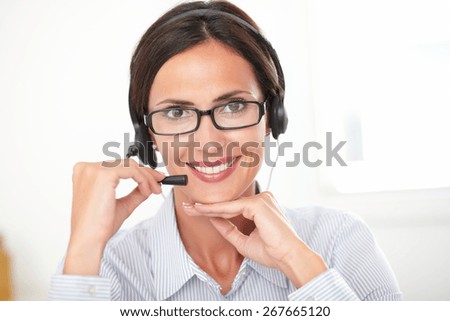 Hispanic receptionist in blue blouse conversing on her headphones while looking at you and smiling cheerfully