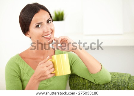 Beautiful young woman toothy smiling at the camera while touching her chin and holding a yellow mug