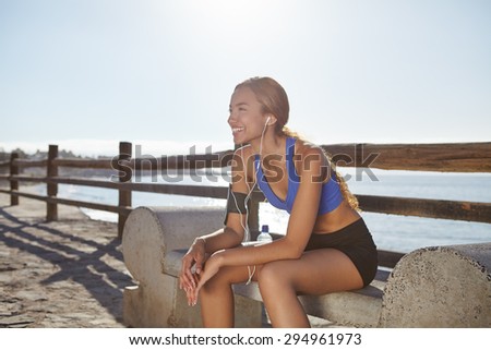 Young female jogger resting on the coastline while listening to music and smiling