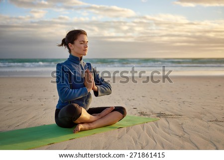 Mature woman sitting in a cross-legged yoga pose on a deserted beach at daybreak, with her hands together in front of her chest, she is focused with her eyes closed