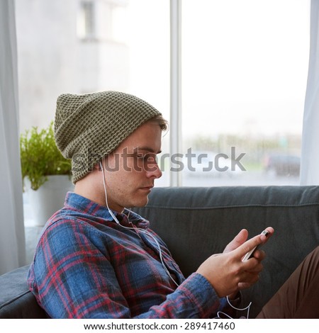 Young guy relaxing on his couch at home with earphones in playing music on his phone