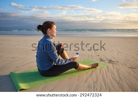 Peaceful woman in sports wear sitting quietly on her yoga-mat on the beach watching the sunrise