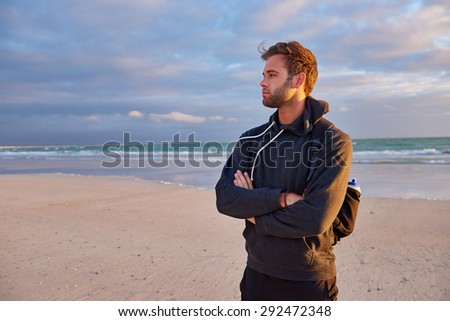 Young guy with his arms crossed looking thoughtfully into the distance while standing on a beach at sunrise