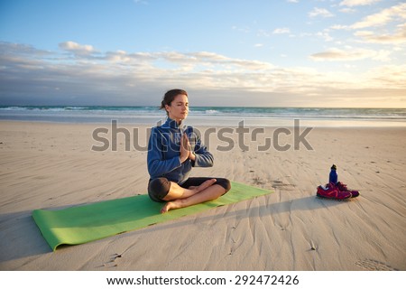 Young woman sitting cross-legged on her yoga-mat on the beach meditating at sunrise