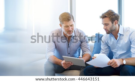 Photo of Mature businessman using a digital tablet to discuss information with a younger colleague in a modern business lounge