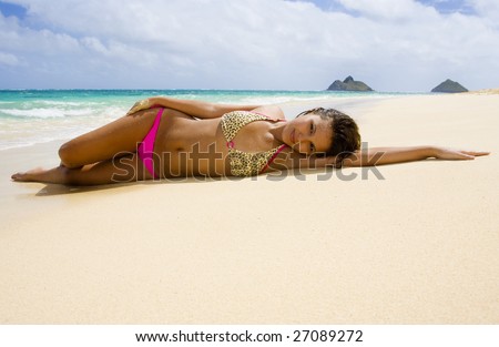 a beautiful young Polynesian girl in a pink bikini lying on a Hawaii beach at water\'s edge with a flower in her hair