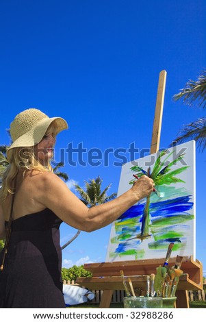 A female artist in her fifties creating a painting outside on canvas of a palm tree and the ocean in Lanikai, Hawaii