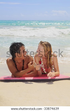 A young Hawaiian man and a blond girl visiting the islands lie on a boogie board by the ocean at the beach in Kailua talking to each other