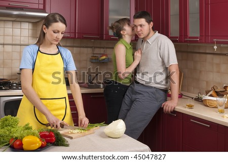 Alone and sad young woman to cut the vegetables in the kitchen. Behind her, two young people are gossiping about her