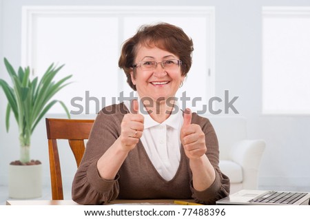 Senior woman showing thumbs up sitting on desk at home