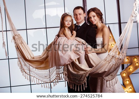 A happy family concept. Beautiful pregnant wife in wedding dress,  her cute daughter and young father are standing near a hammock at a window background.