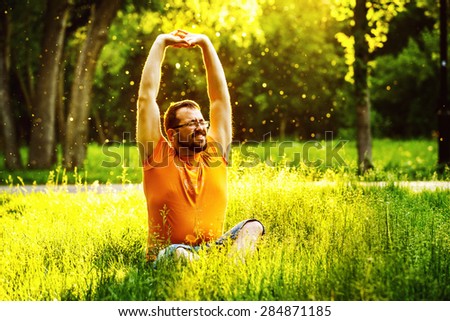 A happy man is stretching himself on green grass with squint eyes and raised up to sky arms at sunny summer day at park background. Concept of wellbeing and healthy lifestyle