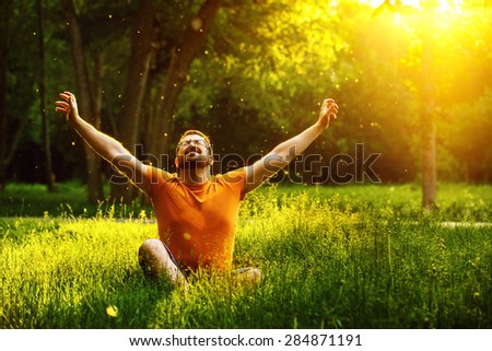 A happy man is relaxing on green grass with squint eyes and raised up to sky arms at sunny summer day at park background. Concept of wellbeing and healthy lifestyle