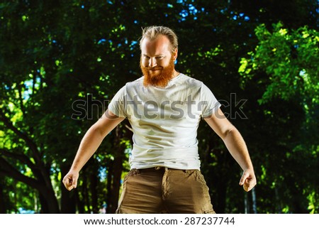 Macho man with red beard is smiling and putting hands down showing attraction at green summer outdoor background.