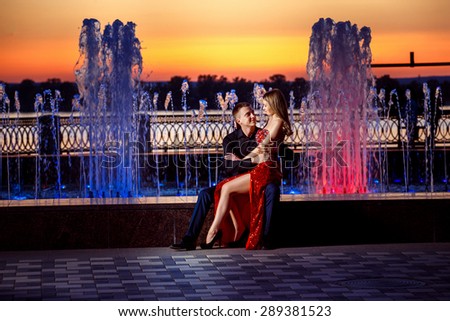 Beautiful young happy couple sitting at colored water fountain and looking at each other at golden stunning sunset background.