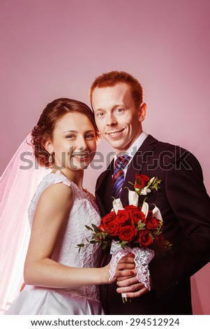 Vertical portrait of happy smiling wedding couple holding red roses flowers bouquet at pink wall background.