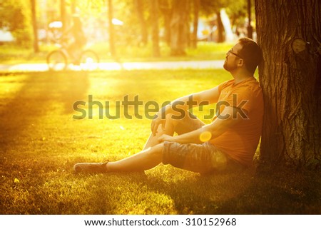 A happy thoughtful dreamer man is sitting on green grass in a park at sunny summer day and looking into future. Concept of relaxation, wellbeing, lifestyle.