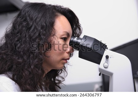 Female chinese scientist sitting at microscope gazing into eyepieces