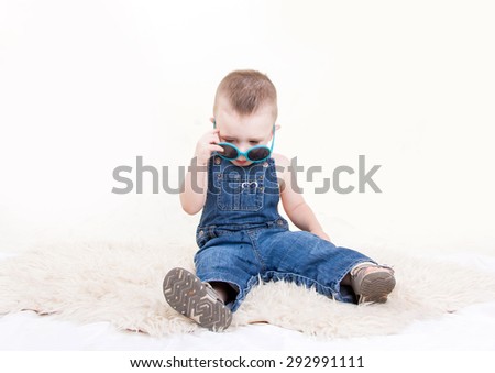 Cute kid in denim overalls with sunglasses sitting isolated on white background
