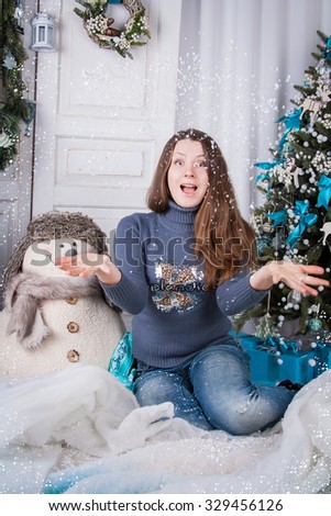 Young beautiful  girl is happy new year holiday. A girl sits among Christmas trees and snowmen, and happily throwing up hands artificial snow. New Year - the most cheerful and desirable holiday